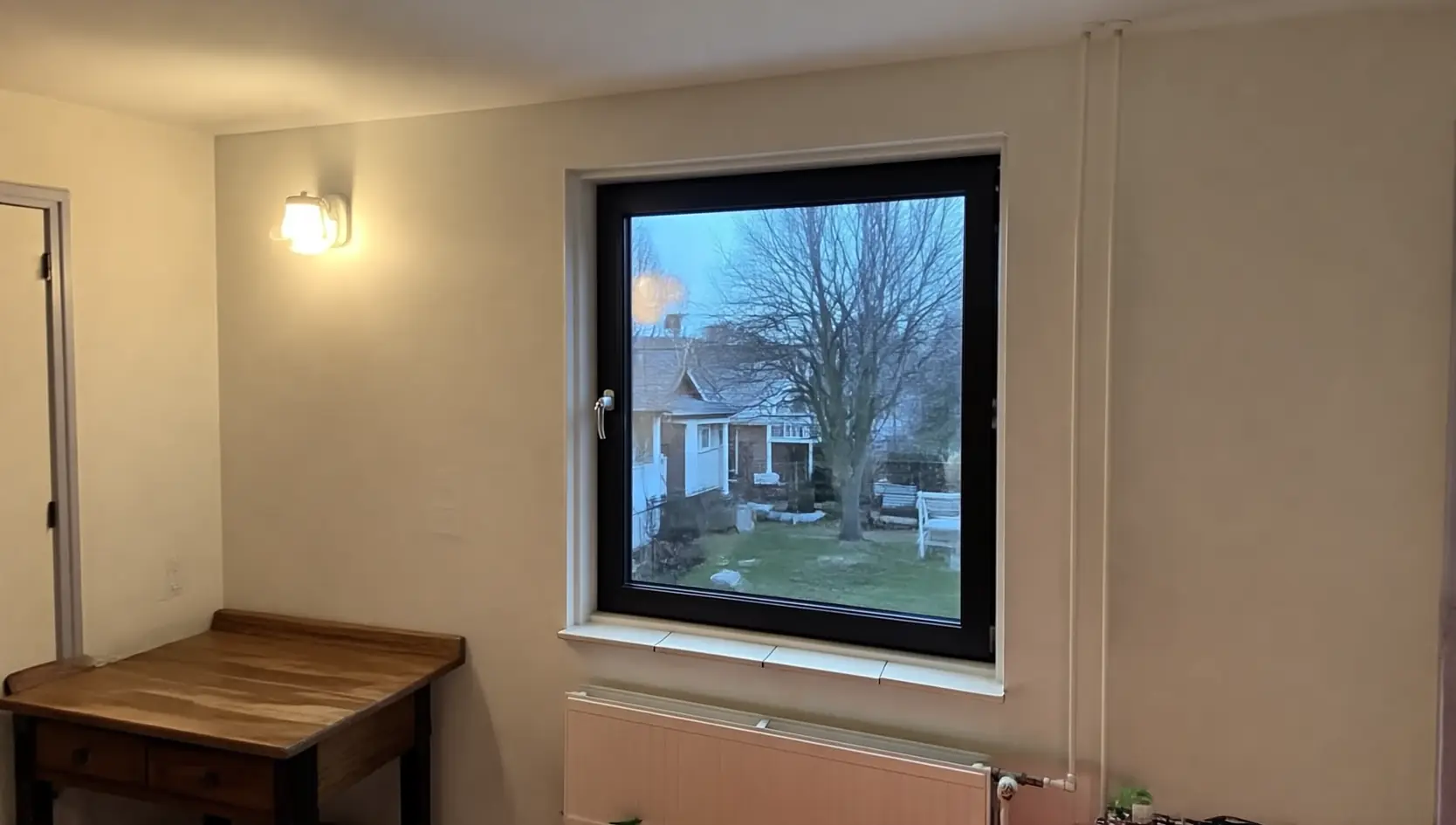 Zwart kunststof draairaam in een lichte binnenwand boven een radiator, met uitzicht op een tuin in de schemer