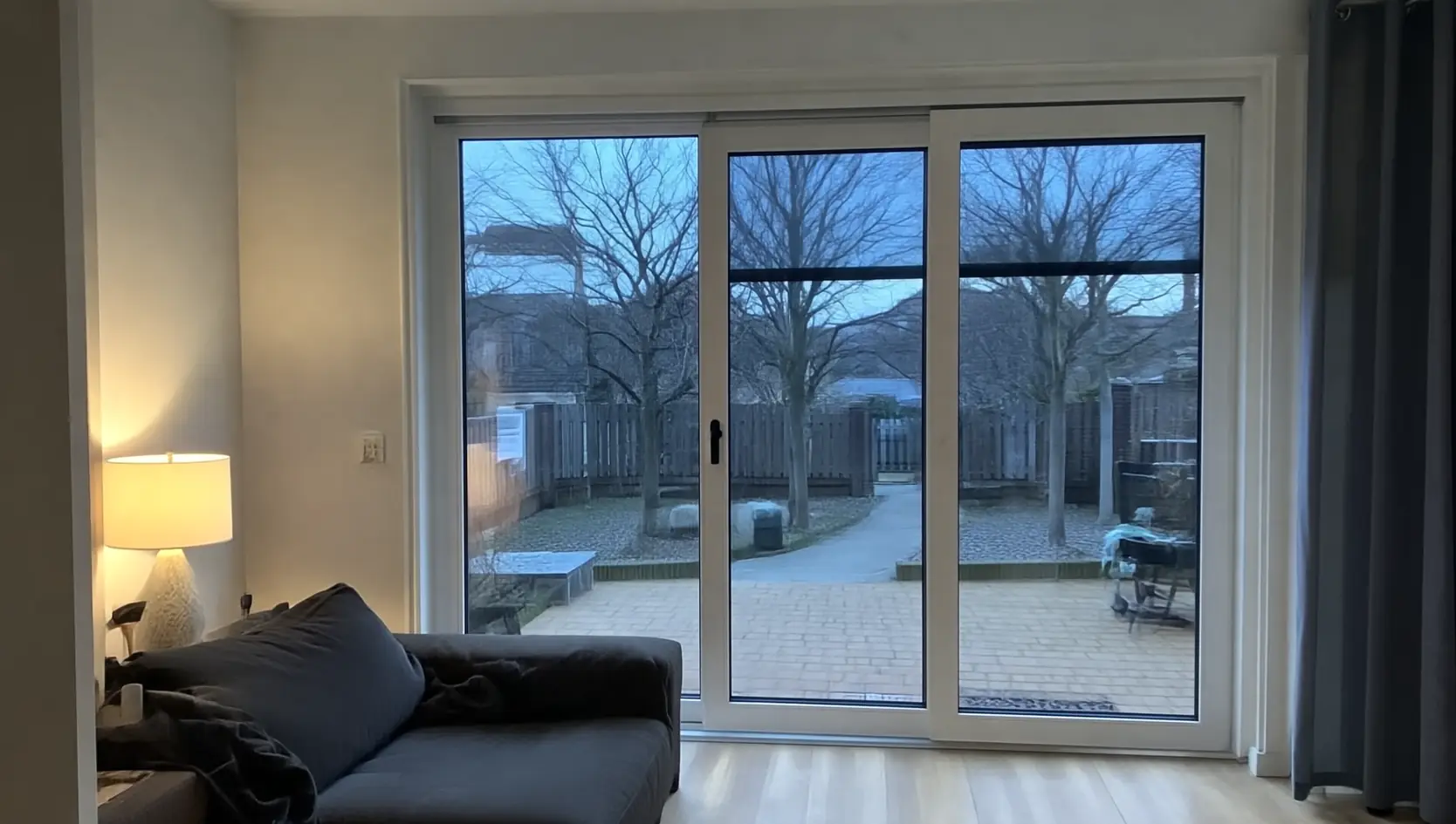 Witte kunststof schuifpui met drie glasvlakken achter een donkere bank, met zicht op een terras bij schemer