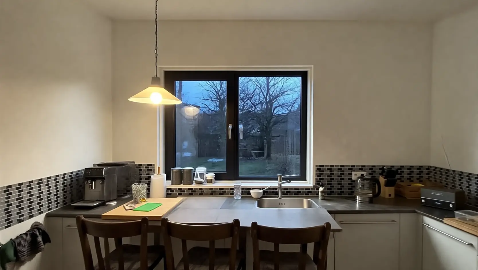 Donker kunststof raam boven het aanrecht in een keuken met hanglamp en eettafel, met zicht op kale bomen bij schemer