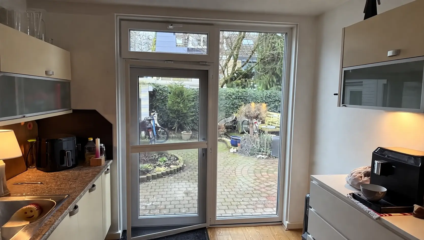 Witte deur met bovenlicht en een vast glazen zijlicht in een keuken, met zicht op een bestrate tuin met fietsen, bij daglicht