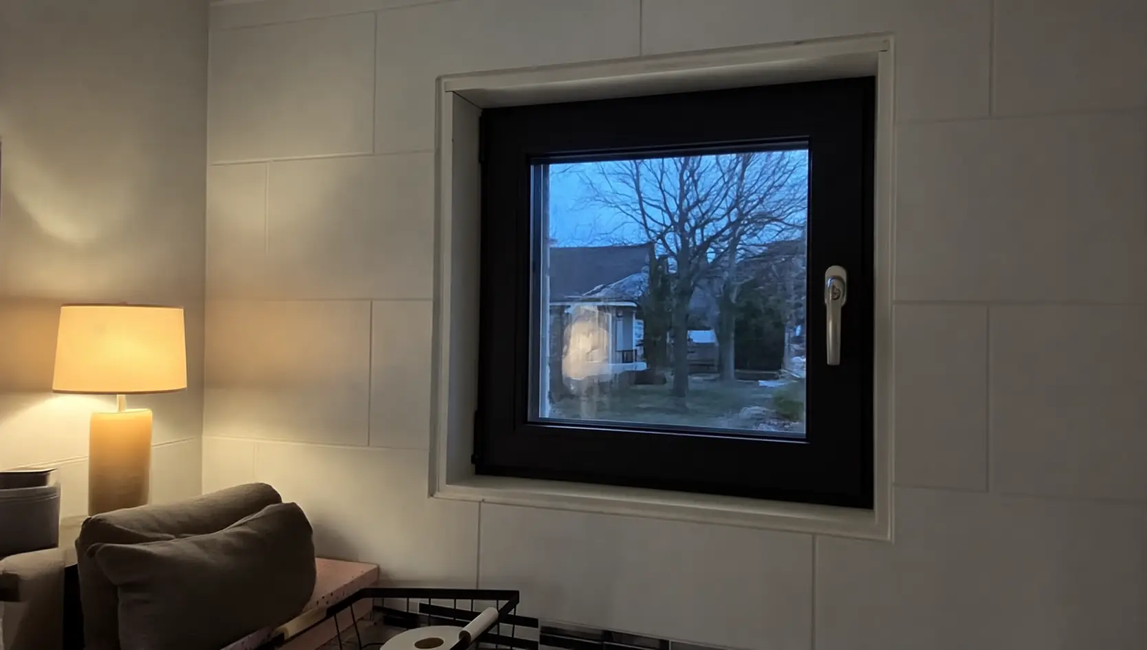 Zwart draaikiepraam met kruk in een lichte wand naast een staande lamp, bij schemer