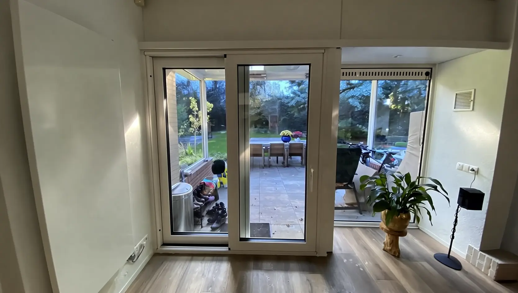 Witte schuifdeur met twee glasvlakken in een kamer met een plant, met zicht op een overdekt terras met tuintafel en een tuin, bij daglicht
