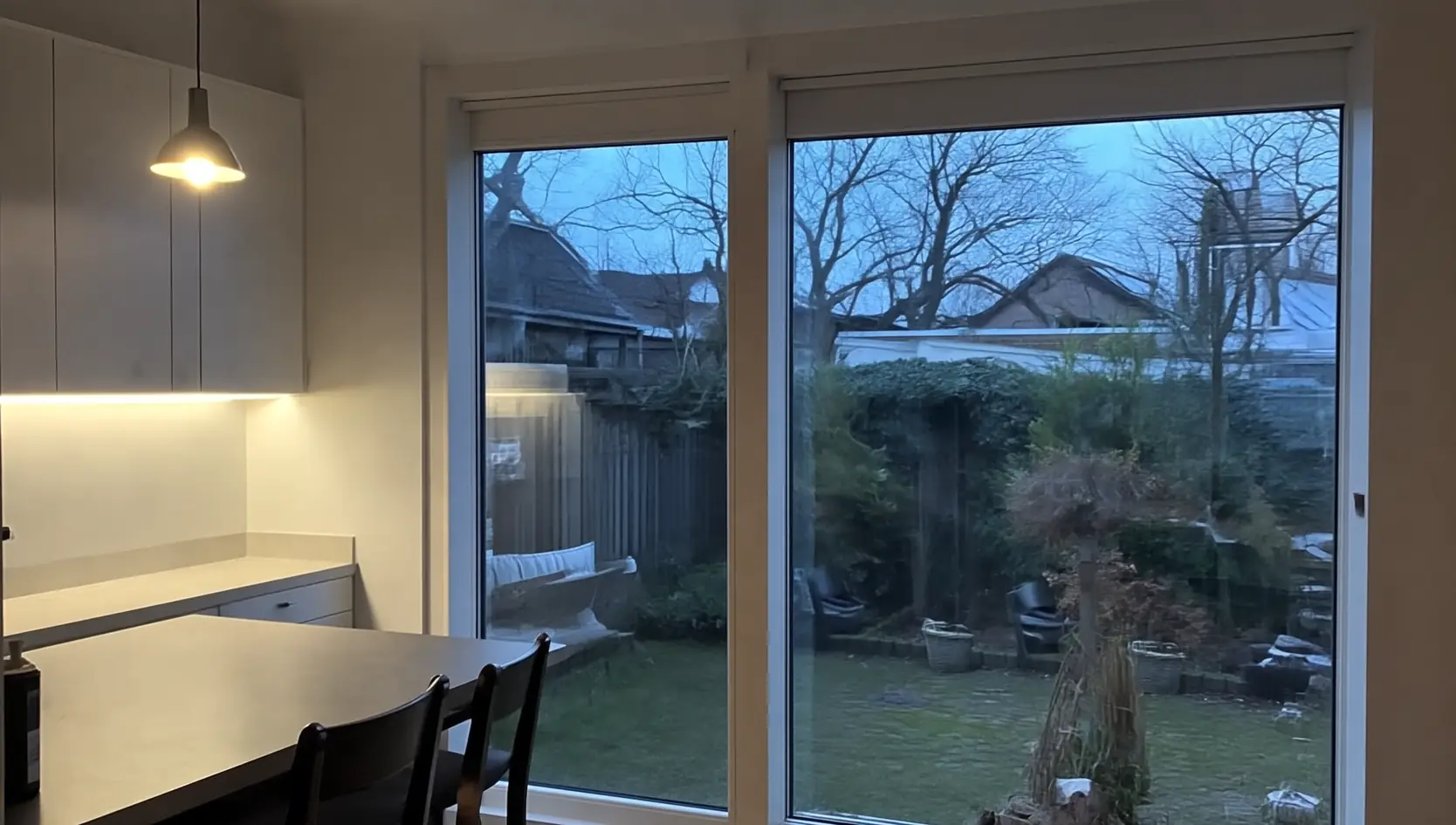 Wit kozijn met twee brede glasvlakken tot de vloer in een keuken-eetkamer met hanglamp boven de tafel, met zicht op een tuin met schutting en kale bomen bij avond