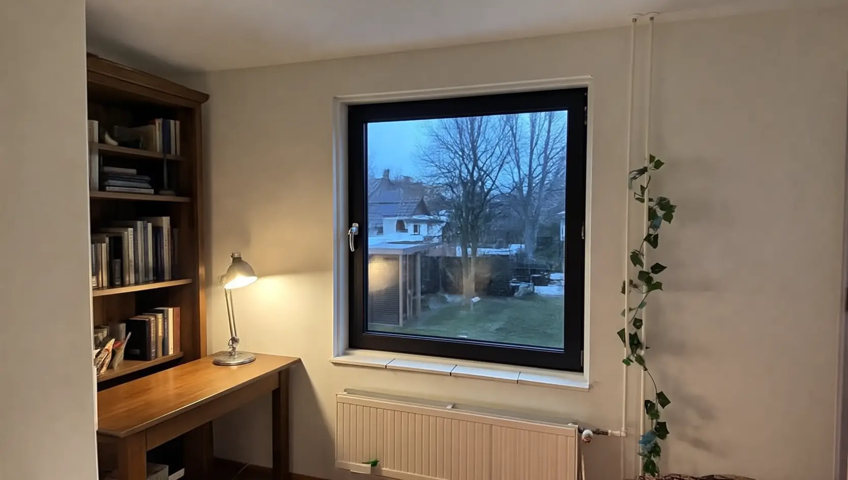 Donker kunststof raam boven een radiator in een werkkamer met boekenkast en bureaulamp, met zicht op een tuin bij schemer