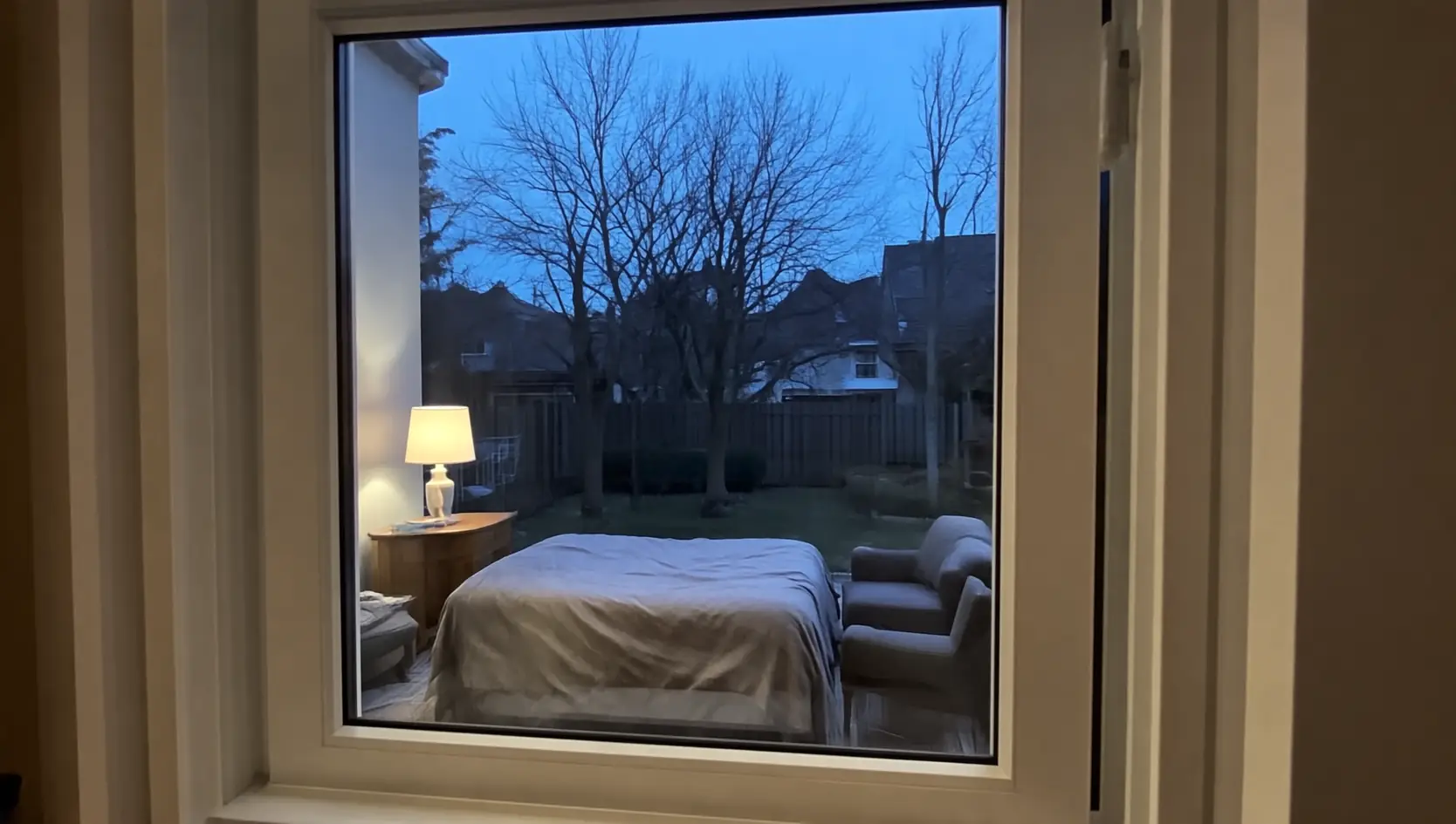 Wit kunststof raam met brede kaders, met door het glas een slaapkamer met bed en lamp en kale bomen bij schemer
