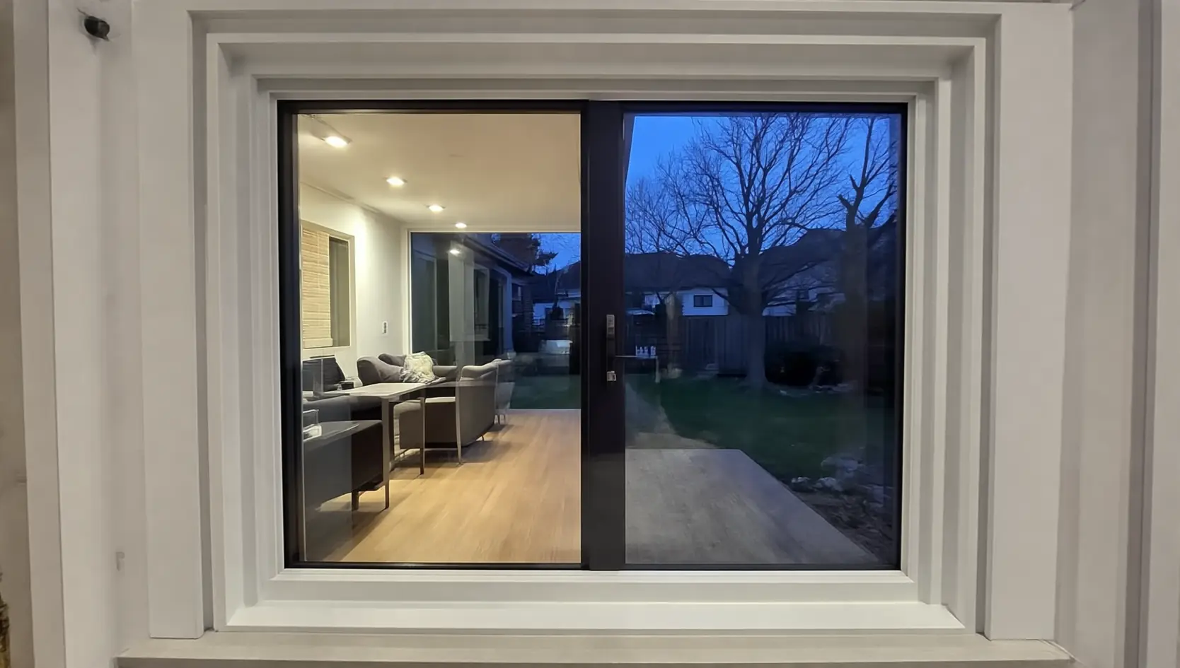 Donker raam met twee glasvlakken in een brede witte omkadering, met door het glas een verlichte zithoek met bank en een tuin met kale bomen bij schemer